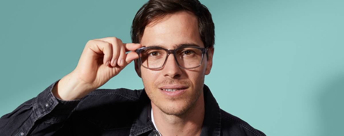 Young man trying on a square pair of glasses in front of a teal wall